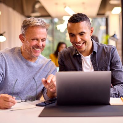 Mature Businessman Mentoring Younger Colleague Working On Laptop At Desk